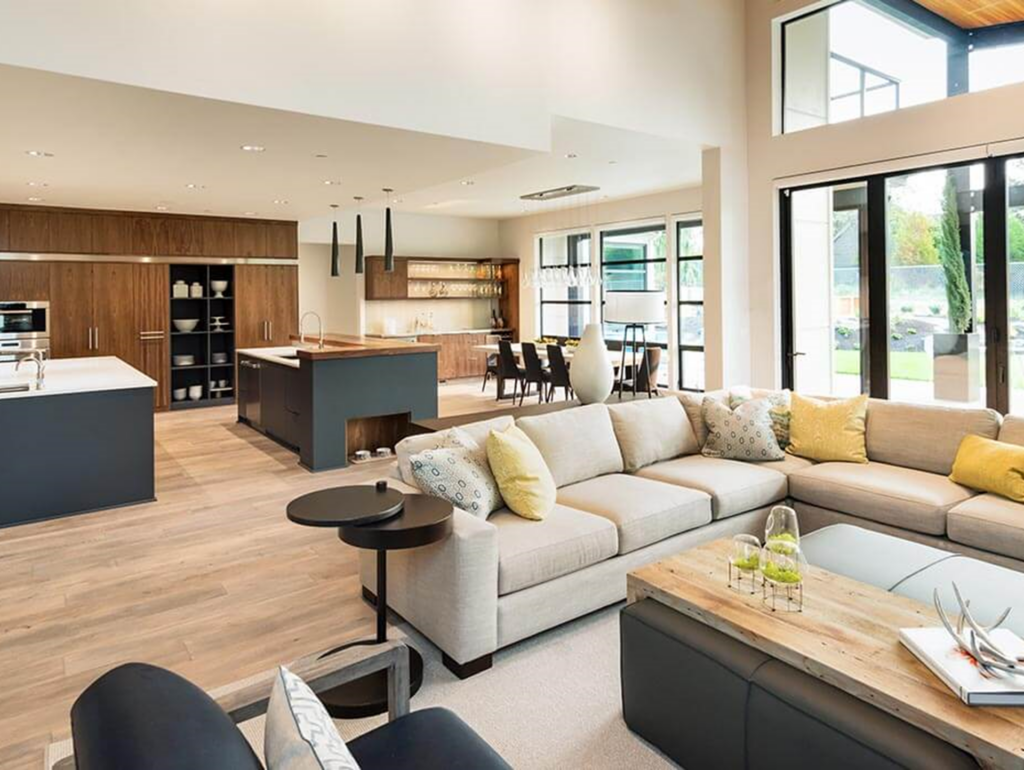 Une vue en angle d'un salon et et fond une salle à manger et une cuisine. bien éclairé avec plusieurs fenestration. Canaper d'une couleur gris pâle avec des cousins jaunes, plancher de la pièces plutôt pâle.